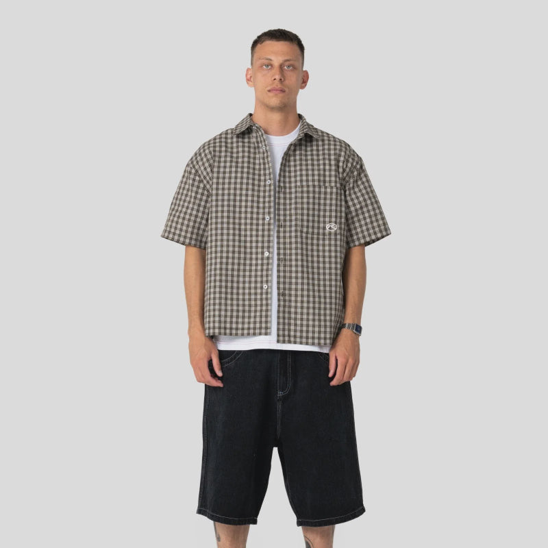A man modeling a short-sleeve brown and beige checkered button-up shirt over a white t-shirt, paired with black shorts, white socks, and black shoes, against a light gray background.