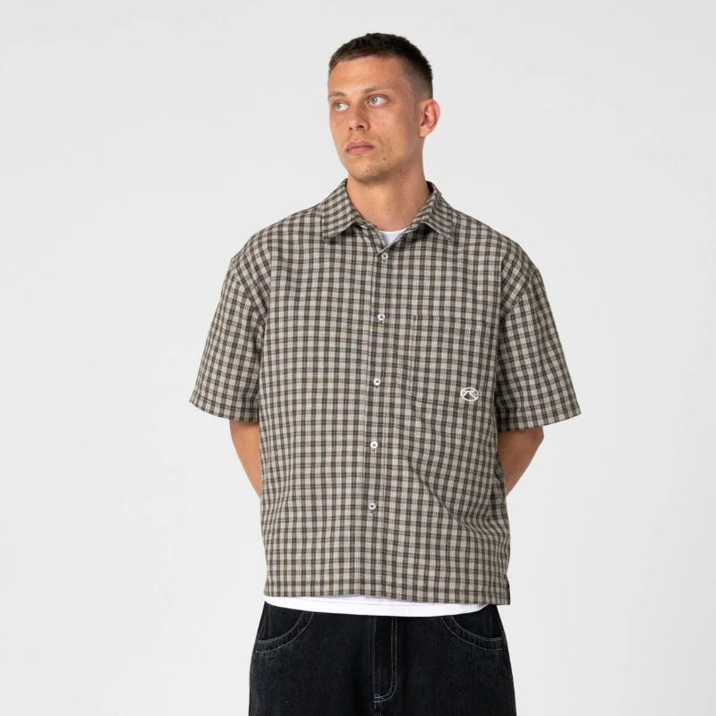 A man modeling a short-sleeve brown and beige checkered button-up shirt, paired with black shorts, white socks, and black sneakers, against a plain light gray background.