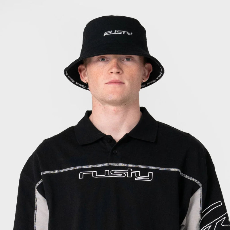 A man wearing a black Rusty brand polo shirt with gray side panels and a matching black bucket hat with a white Rusty logo, photographed against a light gray background.