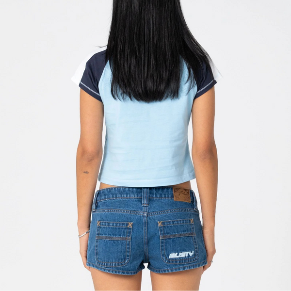 A woman viewed from behind wearing a light blue and navy color-blocked crop top and blue denim shorts against a plain white background.