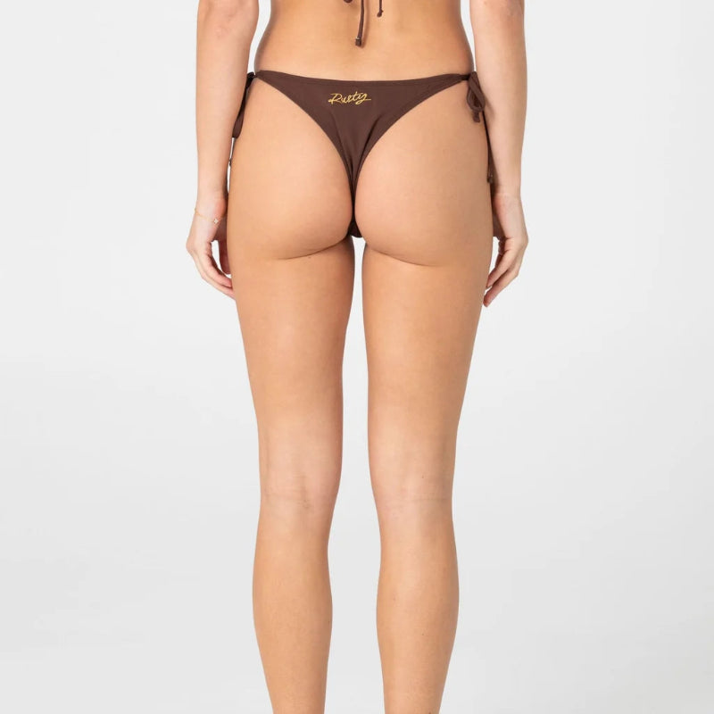 Rear view of a model wearing a brown bikini bottom with side ties and a small gold script logo on the front, photographed against a light gray studio background.