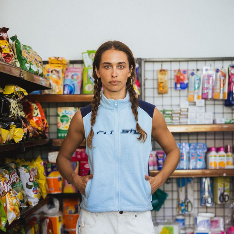A woman with braided hair stands in a retail store aisle wearing a light blue sleeveless zip-up vest with dark navy shoulder panels and white pants.