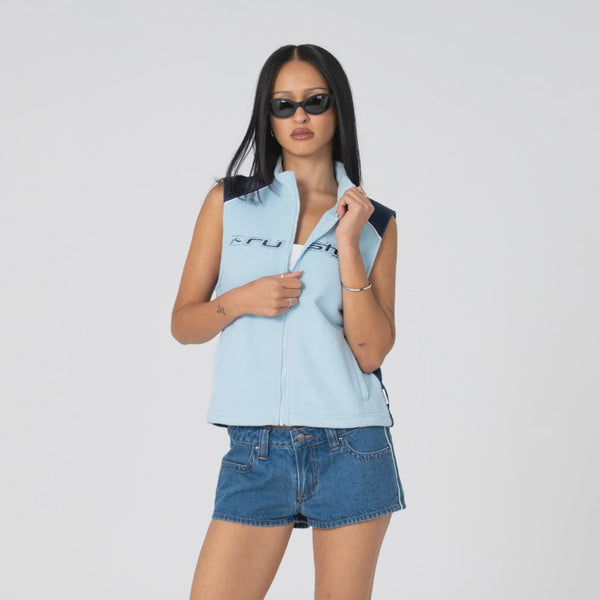 A model wearing a light blue sleeveless zip-up vest with black trim, blue denim shorts, and black slide sandals, standing against a light gray studio background.