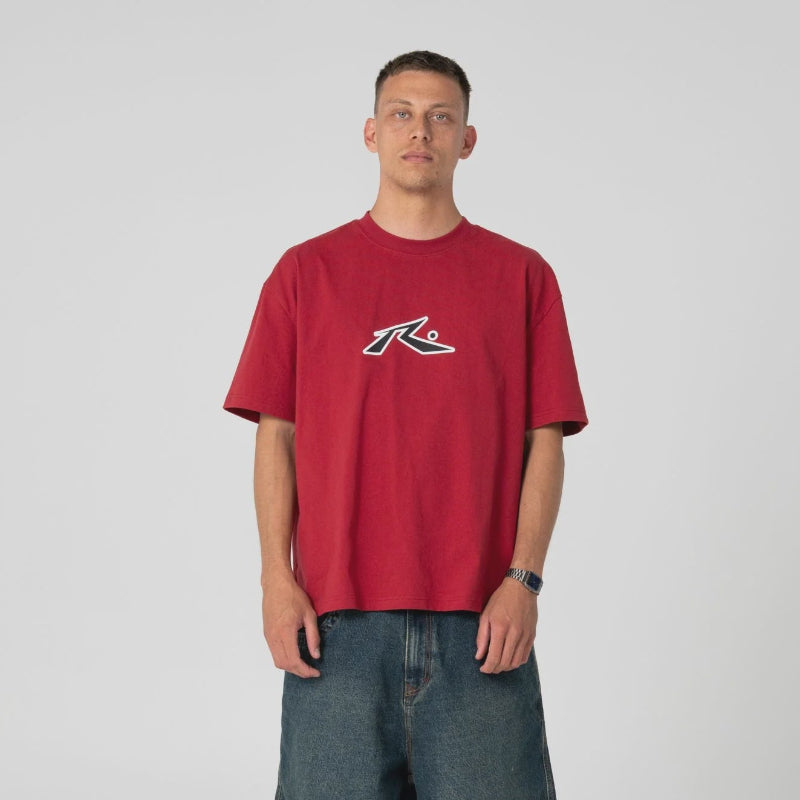 A man modeling a red short-sleeve t-shirt with a white and black graphic logo on the chest, paired with dark blue denim shorts, white socks, and black shoes, against a light gray studio background.