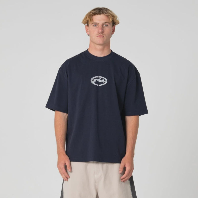 A man stands against a light gray studio background wearing an oversized navy T-shirt with a small white chest logo, light beige shorts with dark side panels, white socks, and dark sneakers.