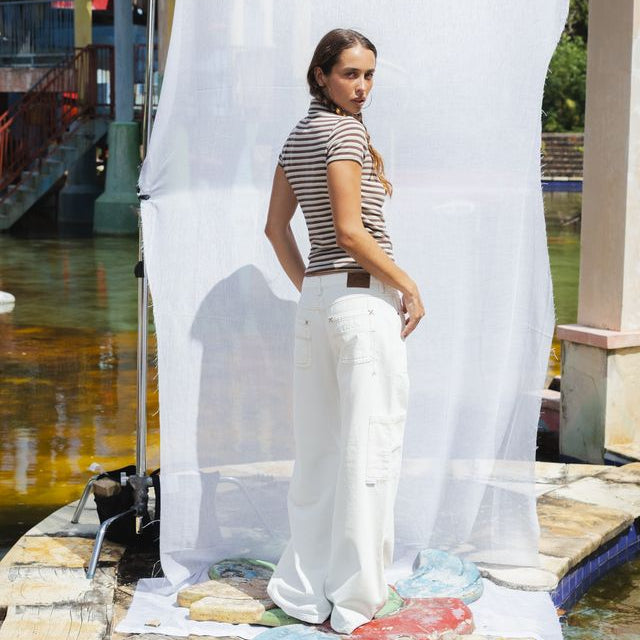 A woman stands in front of a large white fabric backdrop outdoors near a stone walkway and water, wearing a sleeveless black-and-white striped top and white wide-leg pants. Studio lighting equipment is visible to the left, and rocks or fabric samples are scattered on the ground.