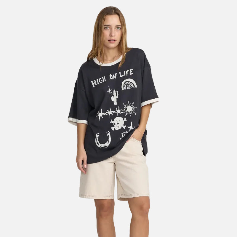 A woman modeling an oversized black graphic T-shirt with white trim and white printed designs, paired with beige shorts and black flats, against a plain white studio background.