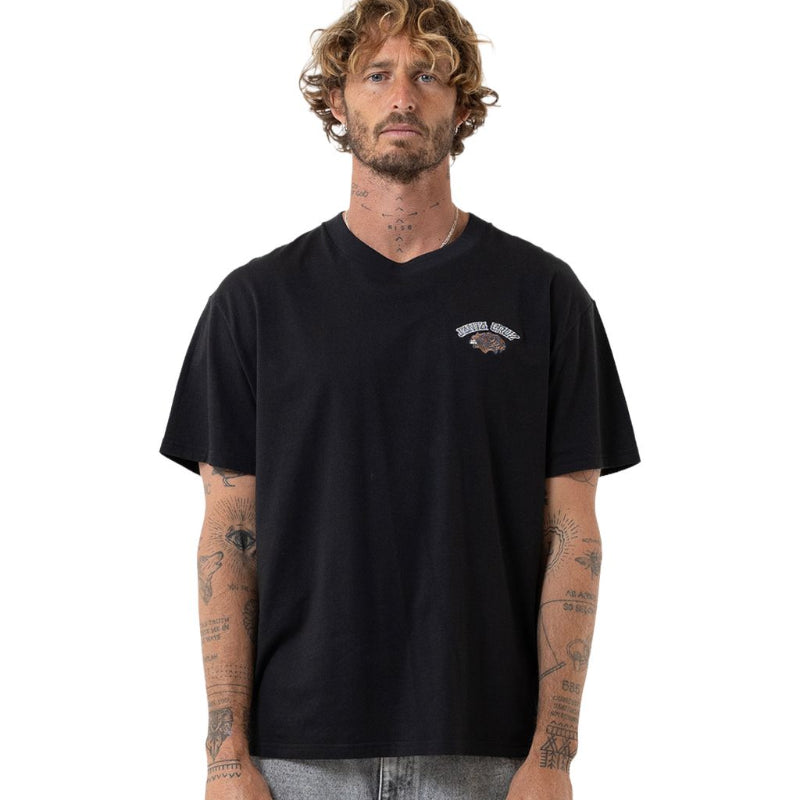A man with curly hair and tattoos wearing a black v-neck t-shirt with a small graphic on the left chest, paired with gray jeans, against a plain white background.
