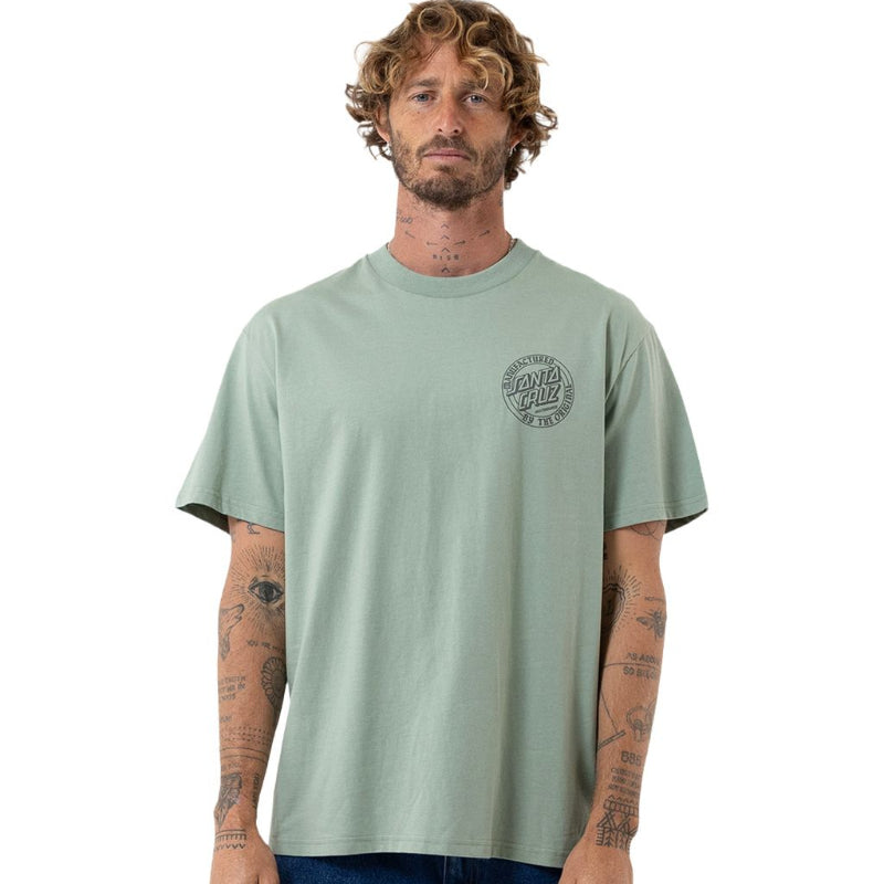 A man wearing a sage green short-sleeve t-shirt with a circular graphic on the left chest, paired with blue jeans, photographed against a solid white background.