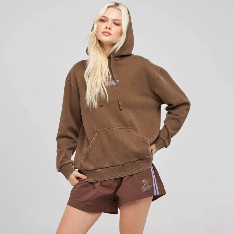 A blonde model wearing a brown washed hoodie, matching brown shorts, white socks, and black Vans sneakers, standing against a light gray studio background.