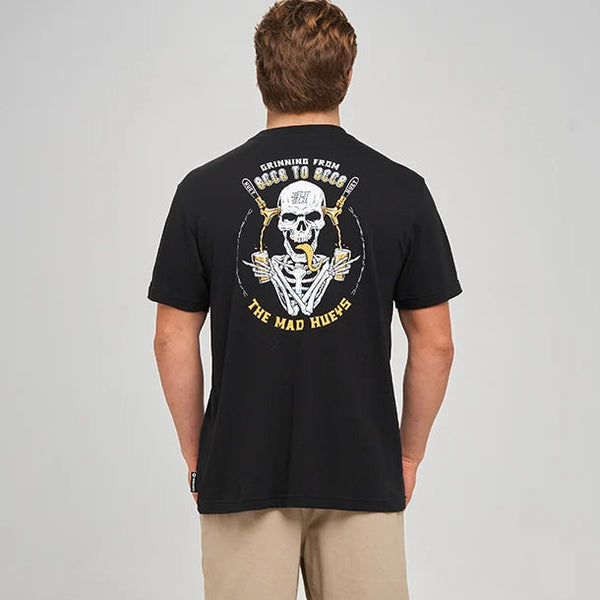 A man viewed from behind wearing a black graphic t-shirt with a large yellow and white skull design on the back, paired with beige shorts, black socks, and white sneakers, against a light gray studio background.