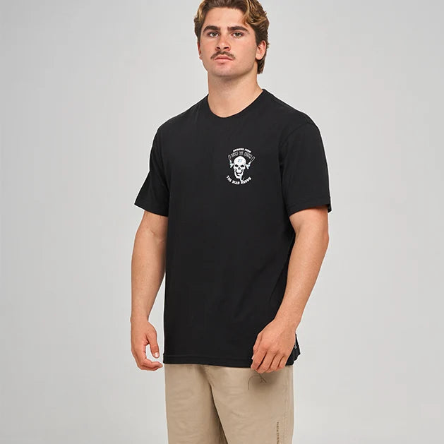 A man modeling a black short-sleeve t-shirt with a small white chest graphic, paired with beige shorts, black socks, and black sneakers, against a light gray studio background.