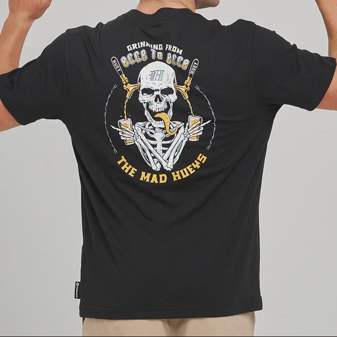 A man shown from the back wearing a black t-shirt with a large graphic on the back. The graphic features a white skull with and beer taps, and the text ' 'THE MAD HUEYS', 'GRINNING FROM BEER TO BEER'