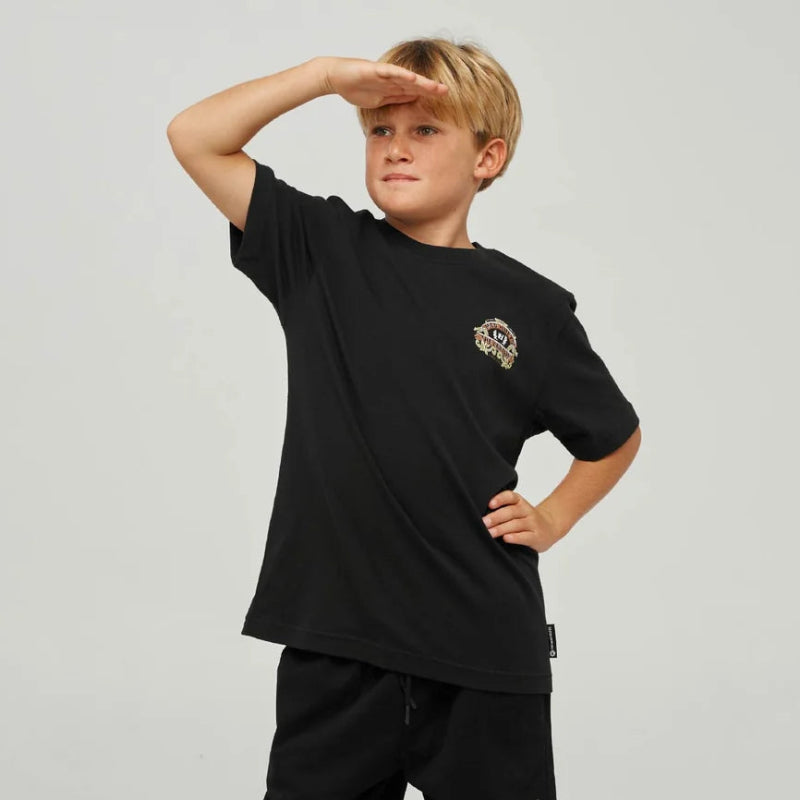 A young boy modeling a black short-sleeve t-shirt with a small graphic on the left chest, paired with black shorts, dark gray socks, and black Vans sneakers, posing against a light gray studio background.