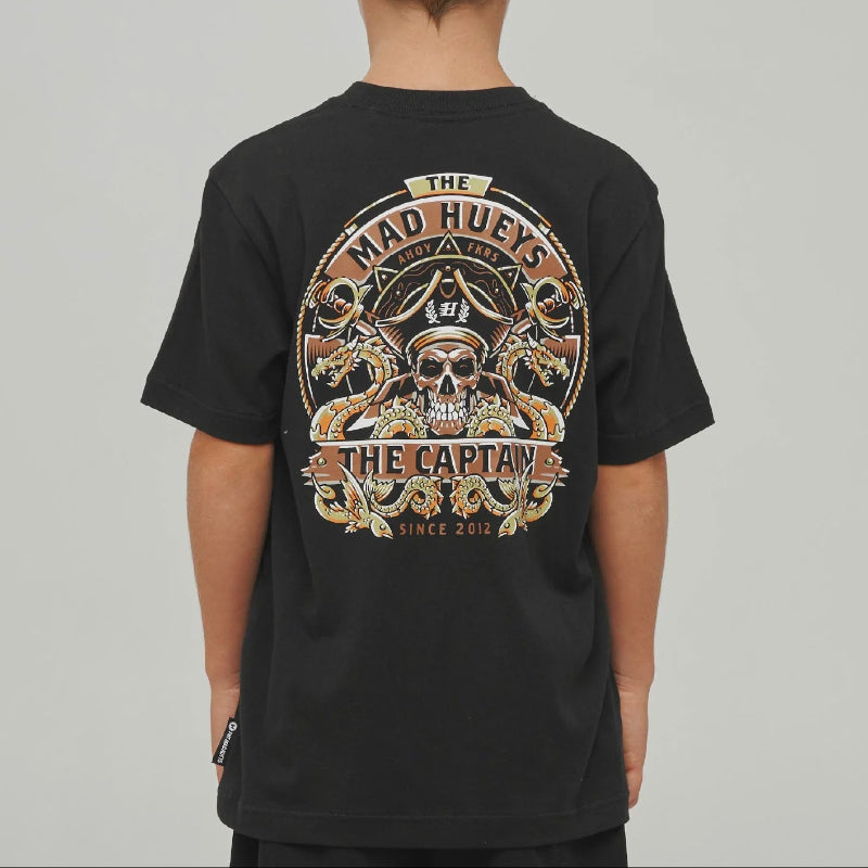 A child viewed from the back wearing a black t-shirt with a large graphic print on the back. The graphic features a skull, pirate hat, and the text 'THE MAD HUEYS  THE CAPTAIN' in a circular design with gold, brown, and white details.