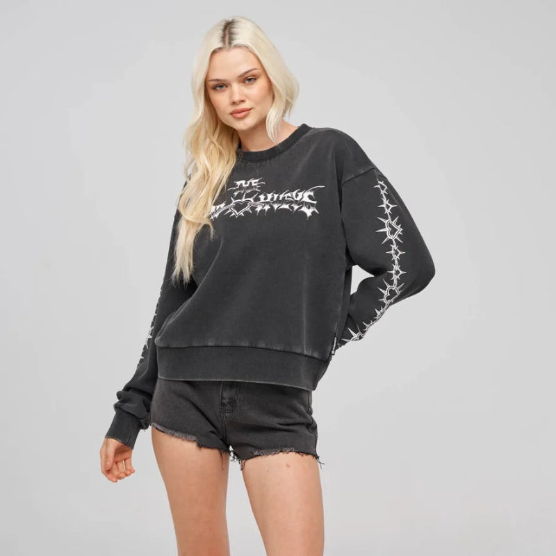 A blonde model wearing a washed black crewneck sweatshirt with white graphic text and barbed wire details on the sleeves, paired with black denim shorts, white socks, and black sneakers, against a light gray studio background.
