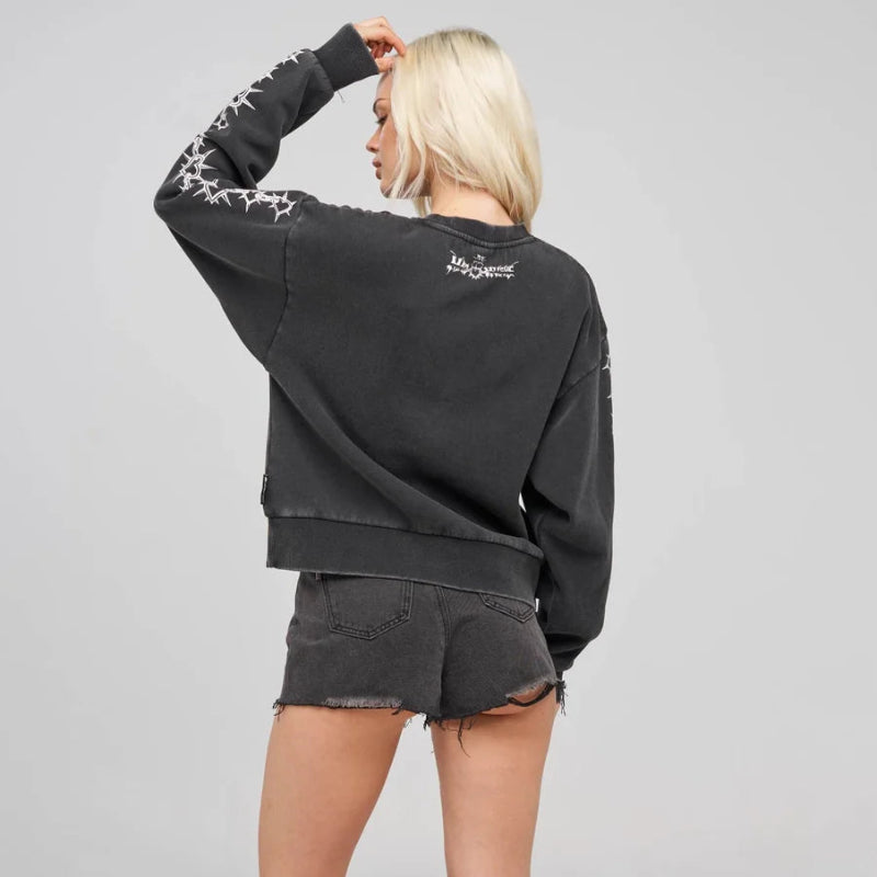 A blonde model shown from the back wearing a faded black crewneck sweatshirt with white graphic prints on the sleeves and back, paired with matching faded black denim shorts, white socks, and black sneakers, against a light gray studio background.