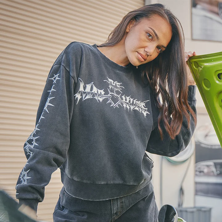A woman wearing a dark gray washed sweatshirt with a white metal-style graphic and thorny trim details, standing next to a bright green car with its hood open.
