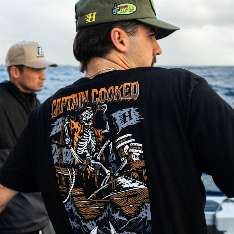 A man on a boat wearing a black Captain Cooked t-shirt with a large skeleton pirate graphic on the back, paired with an olive green cap. Another man is visible in the background.