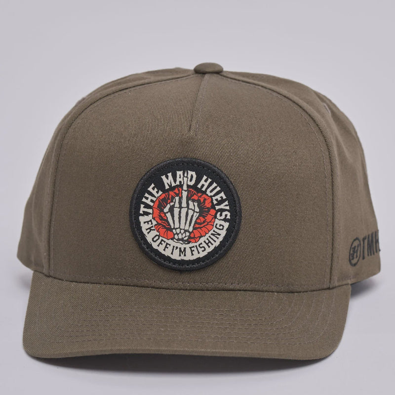 An olive green baseball cap with a circular embroidered patch on the front that reads 'THE MAD HUEYS FK OFF IM FISHING'. The side of the cap features a small black embroidered '@THM' logo.