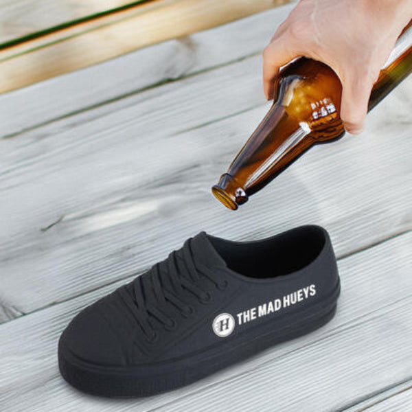 Black sneaker with 'The Mad Hueys' logo on a wooden surface, with a hand holding a bottle.
