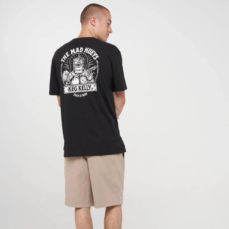A man wearing a black short-sleeve graphic T-shirt with a large white back print, paired with beige shorts, black socks, and black sneakers, photographed against a light gray studio background.