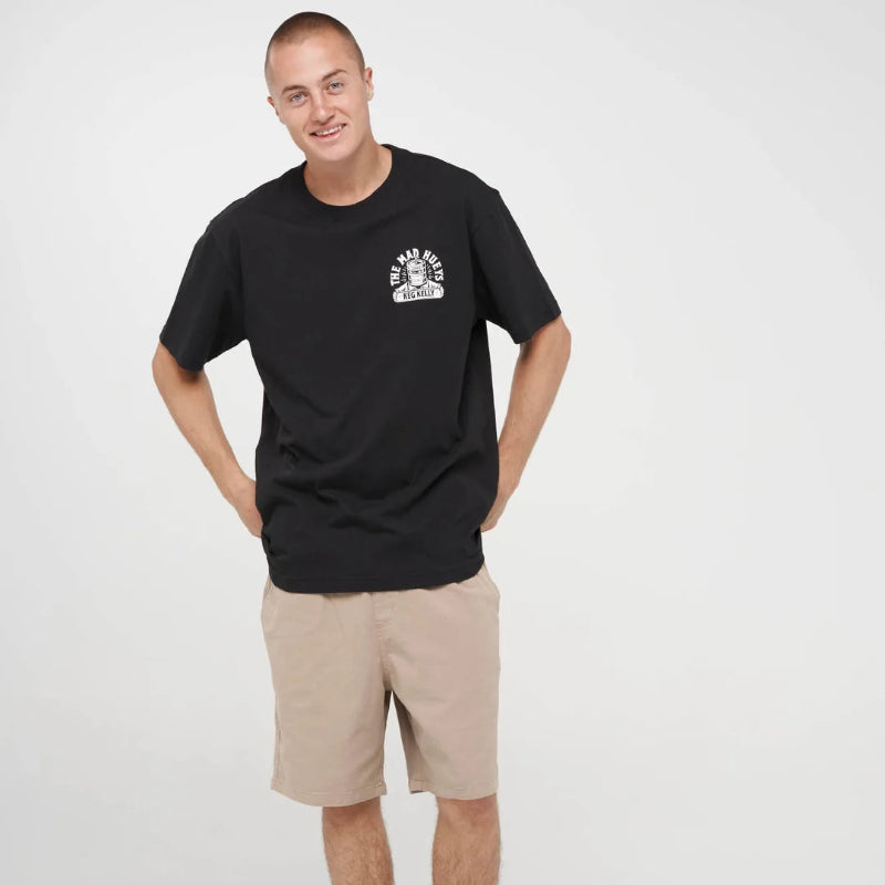 A man modeling a black crewneck t-shirt with a small white graphic on the left chest, paired with beige shorts, black socks, and black sneakers, against a plain white studio background.