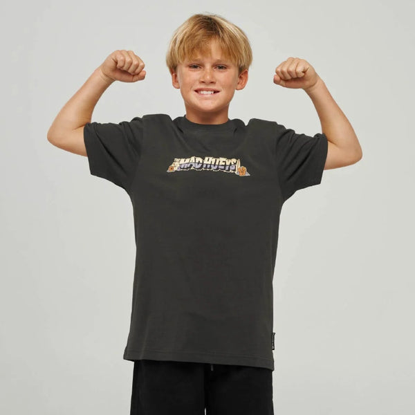 A young boy posing in a black short-sleeve t-shirt with a colorful graphic on the chest, paired with black shorts, gray socks, and black sneakers, against a light gray studio background.