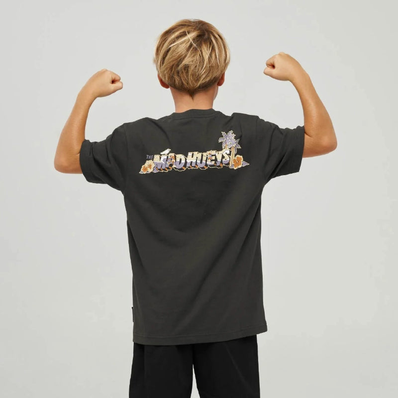 A child model shown from the back wearing a black graphic t-shirt with 'MAD HUEYS' printed across the upper back, paired with black shorts, gray socks, and black sneakers, against a light gray studio background.