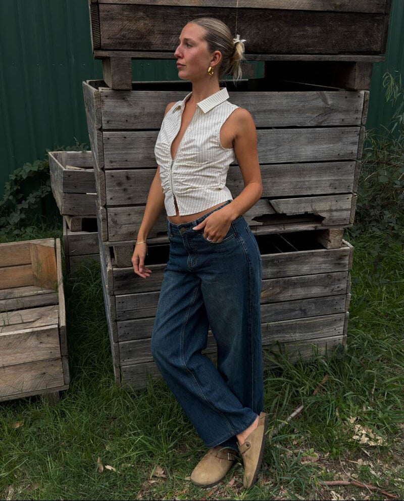 Woman standing in front of wooden crates wearing a white sleeveless shirt and blue jeans.