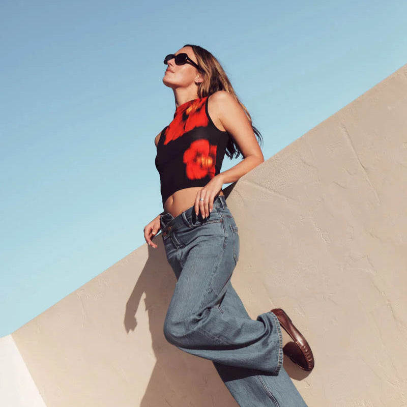Woman in a red floral top and blue jeans leaning against a sand dune with a clear blue sky.