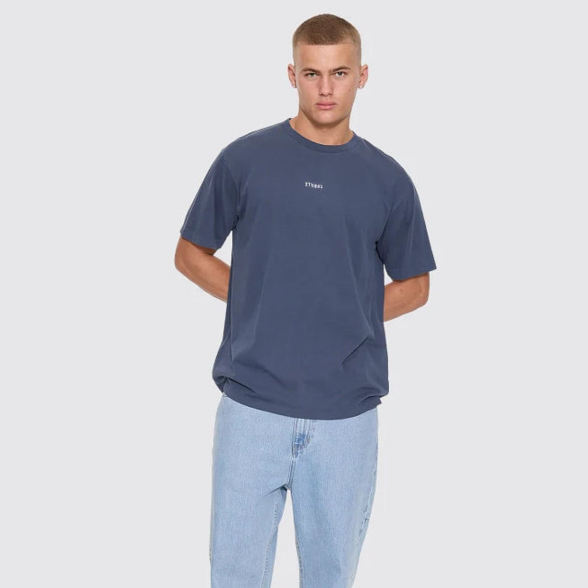 A male model wearing a slate blue crewneck t-shirt with a small white logo on the chest, paired with light wash jeans and black slip-on sneakers, standing against a solid light gray background.