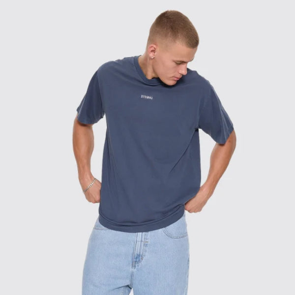 A man wearing a navy blue crewneck t-shirt with a small white logo on the chest, paired with light wash blue jeans and black slip-on shoes, standing against a light gray studio background.