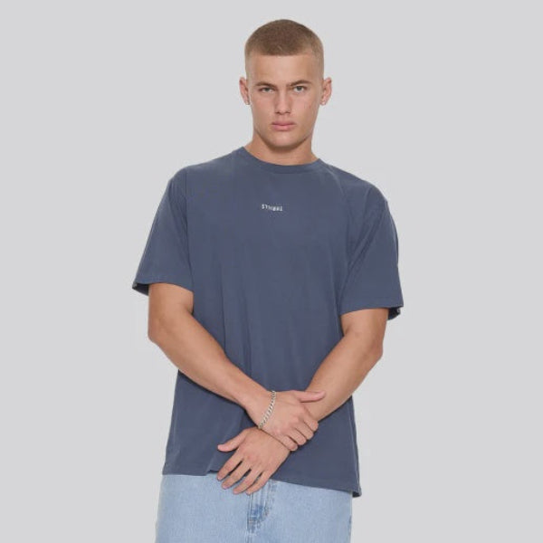 A male model wearing a dark slate blue t-shirt, light wash jeans, and black slip-on shoes, standing against a solid gray background.