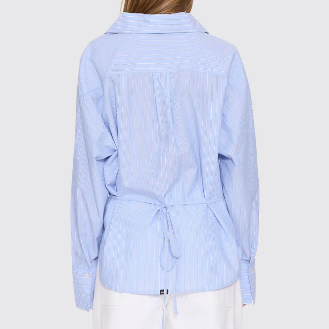 A woman viewed from the back wearing a light blue and white vertically striped long-sleeve shirt with a tie waist, paired with white pants, against a light gray studio background.