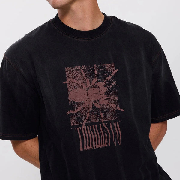 A man wearing a black crewneck t-shirt with a distressed dusty rose graphic of a spider and the word 'THRILLS CO' printed on the front, photographed against a light gray background.