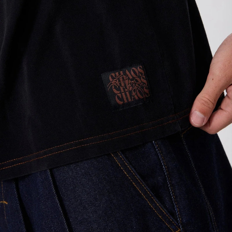 Close-up of a person wearing a black oversized T-shirt with a visible 'Chaos Chaos Chaos' brand patch on the lower front, paired with dark blue jeans against a plain light background.
