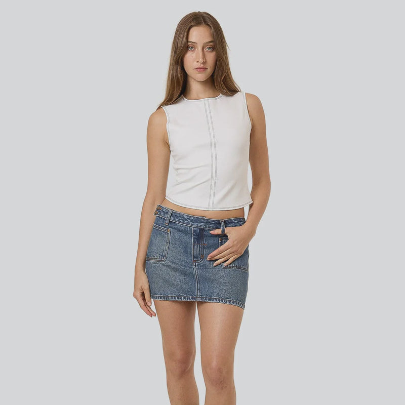 Woman wearing a white sleeveless top and denim skirt with black boots on a plain background