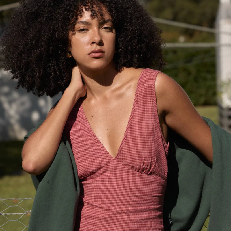 A woman with a large dark curly afro stands outdoors wearing a sleeveless deep V-neck ribbed dress in a dusty rose pink color, with a dark green cardigan draped over her shoulders.