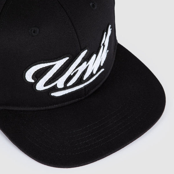 A close-up shot of a black snapback cap featuring a white embroidered script logo on the front panel, set against a light gray background.
