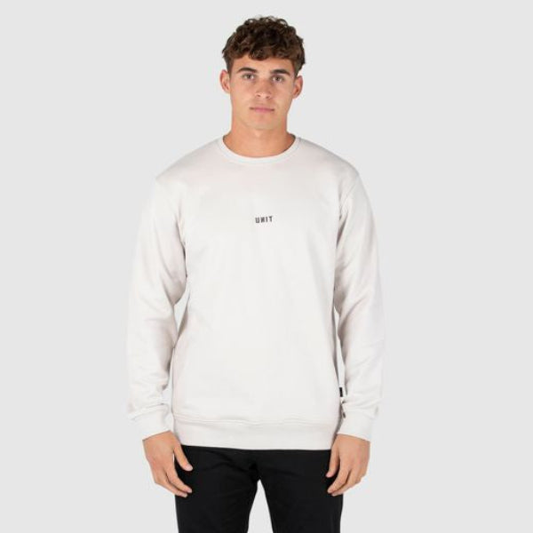 A man modeling a white crewneck sweatshirt with a small black 'UNIT' logo on the left chest, paired with black pants and black shoes, against a light gray studio background.