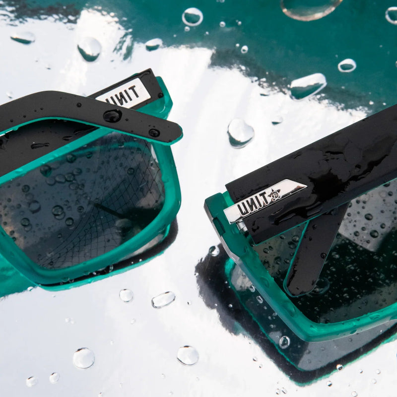Close-up of two teal and black teal sunglasses with water droplets on them, resting on a wet reflective surface. The brand name 'UNIT' is visible on the strap.