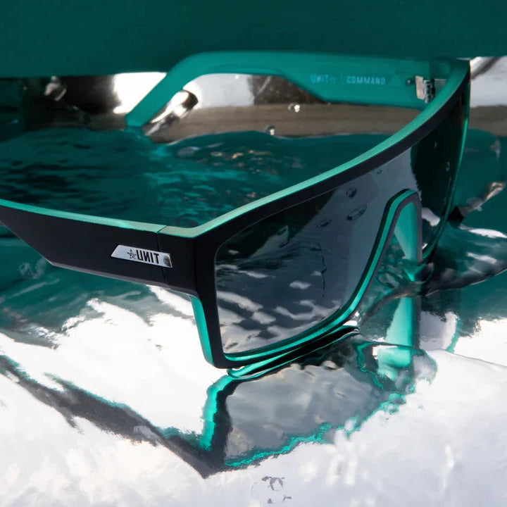 A pair of wraparound sunglasses with a black frame and translucent teal lenses, partially submerged in water on a reflective white surface.