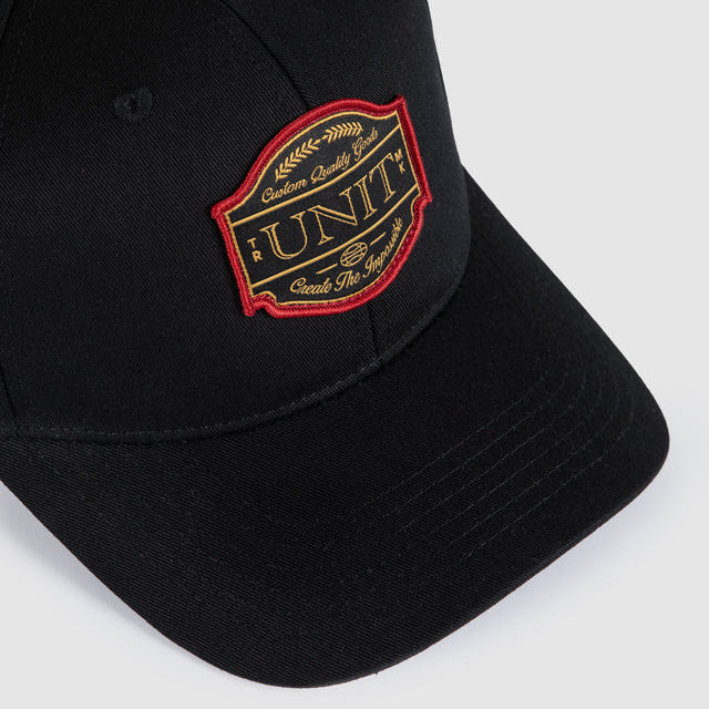 A close-up of a black baseball cap featuring a prominent embroidered shield patch on the front panel. The patch is outlined in red and features gold embroidery with the word 'UNIT' and smaller text reading 'Custom Quality Goods'