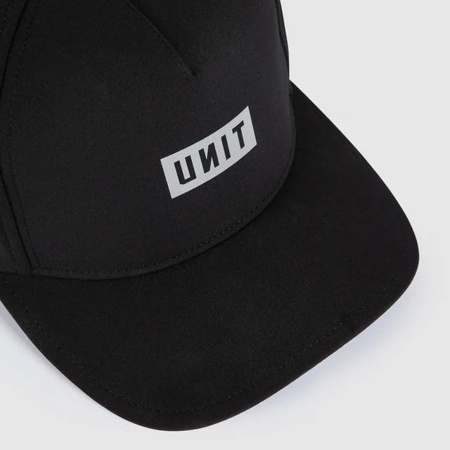 A close-up of a black baseball cap with a white rectangular patch on the front displaying the brand name 'UNIT' in black letters, set against a light gray background.