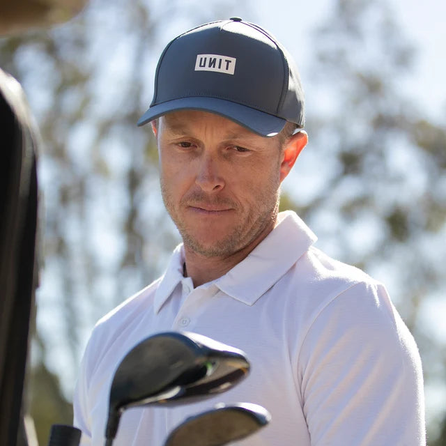 A man wearing a white polo shirt and a dark grey cap with a visible UNIT logo, standing outdoors near golf clubs.