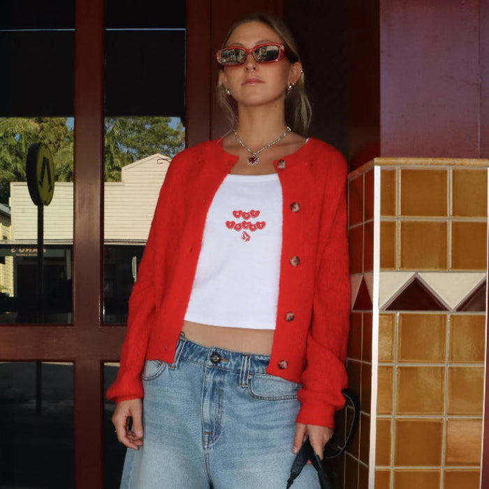 Person wearing a red cardigan, white crop top with text, and blue denim shorts, holding a camera.