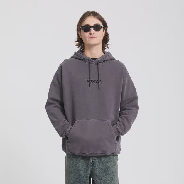 A model wearing a dark gray washed hoodie with 'VOLCOM printed on the chest, paired with dark green acid-wash jeans and black sunglasses, standing against a light gray studio background.