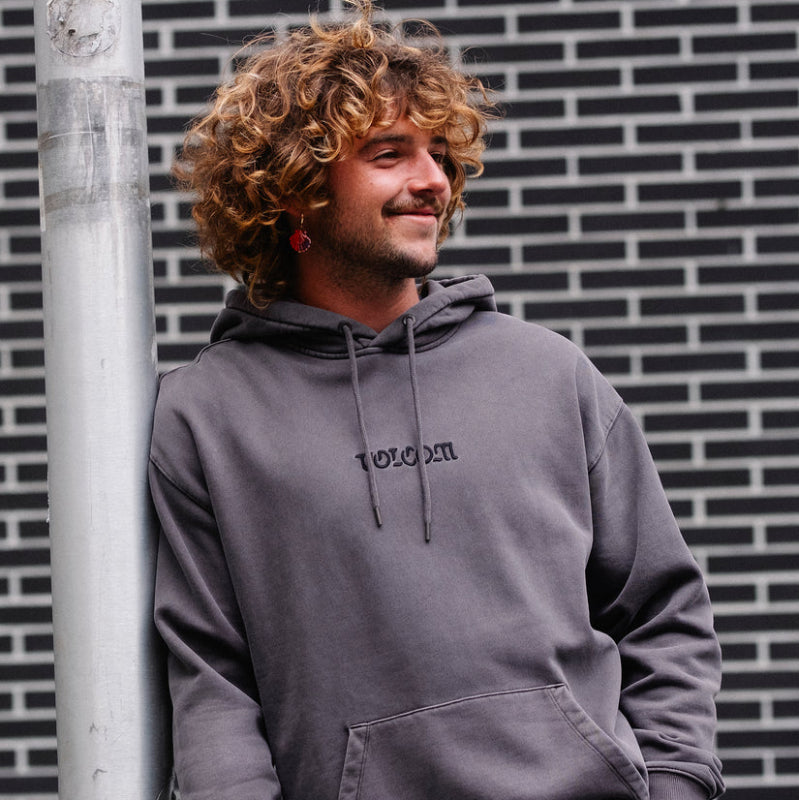 Man wearing a gray hoodie with 'VOLCOM logo against a brick wall.