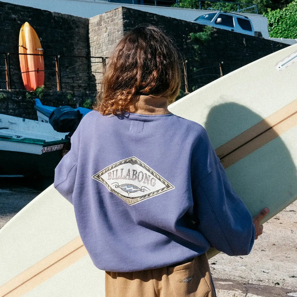 Person holding a surfboard with a Billabong sweatshirt, standing on a stone path.